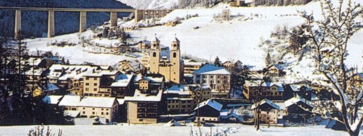 Schneebedeckte Stadt Steinach mit mehreren Gebäuden, darunter zwei Türme im Hintergrund, umgeben von Bergen und Bäumen.